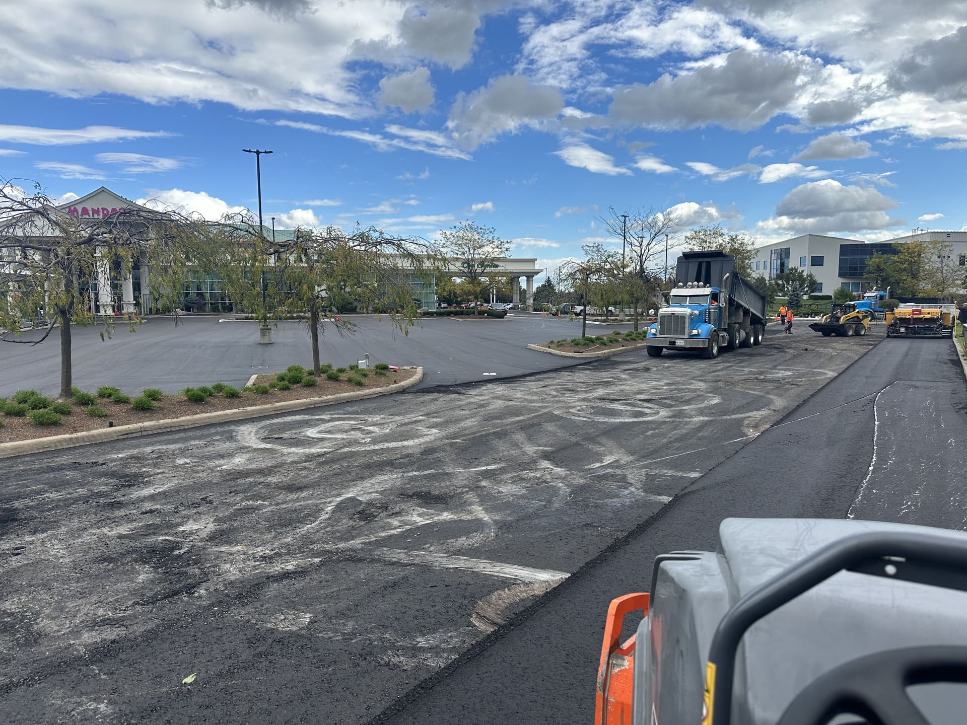 Asphalt repaving underway in a commercial lot with trucks, workers, and freshly laid blacktop under a cloudy sky.