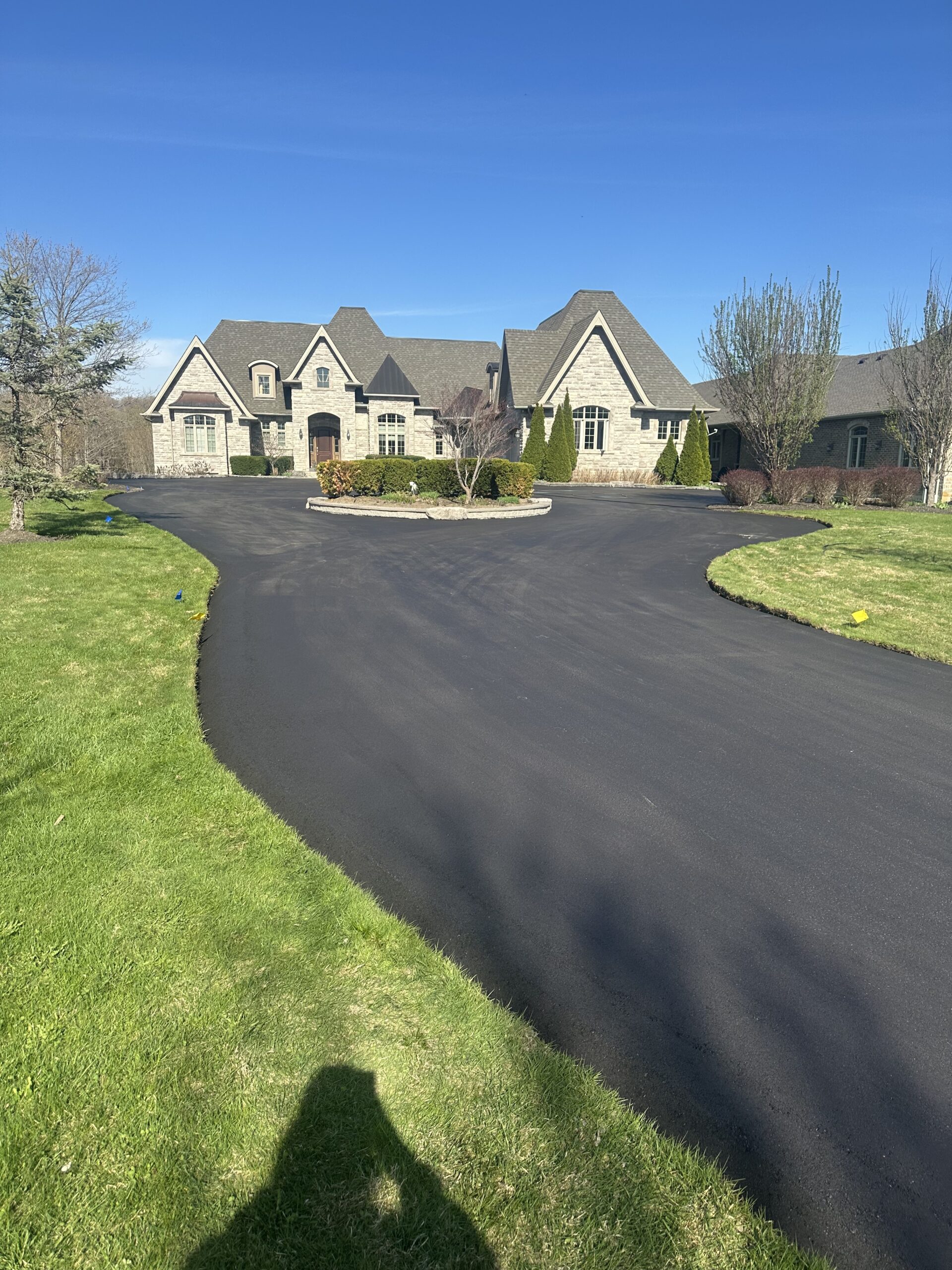 Newly paved residential asphalt driveway leading to a large stone home, surrounded by green lawn and landscaping under a clear blue sky.
