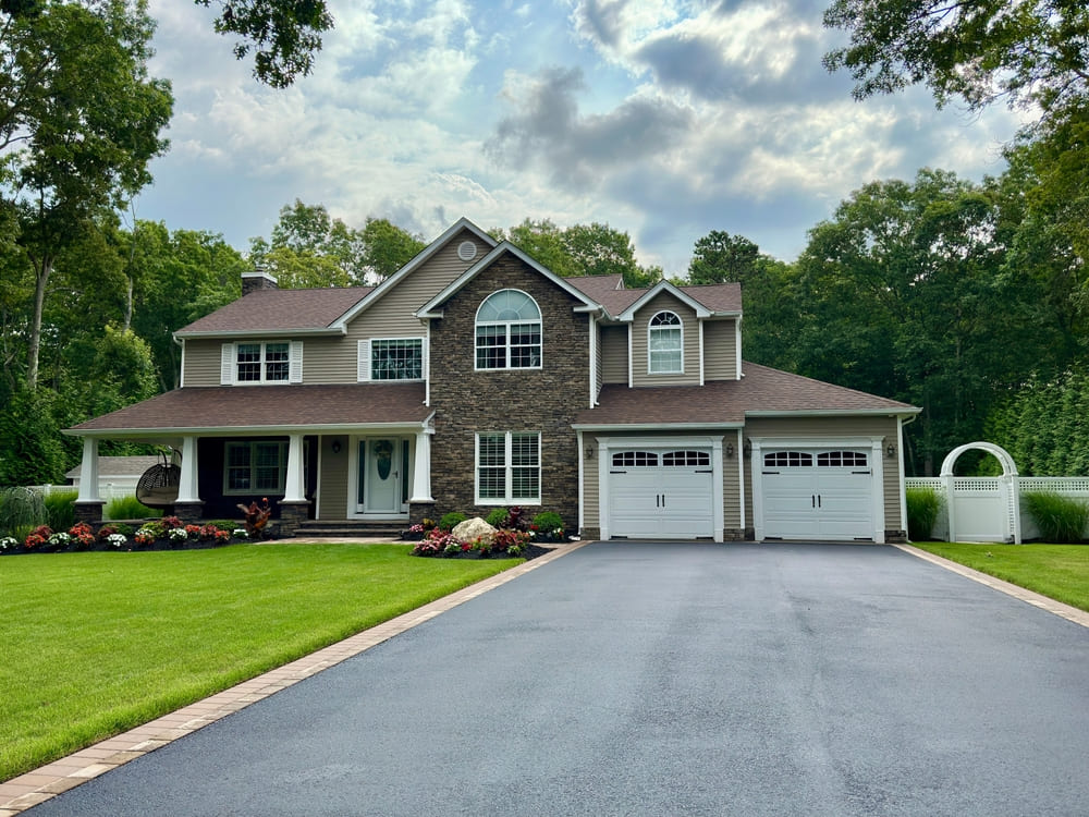 Beautiful suburban home with a freshly paved black asphalt driveway, manicured green lawn, landscaped flower beds, and double garage under a partly cloudy sky.